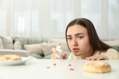 Photo of Emotional young woman spraying insecticide onto cockroaches at white table indoors. Pest problem