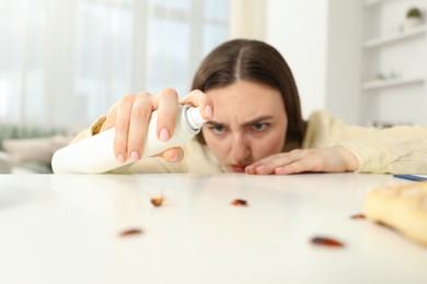 Photo of Emotional young woman spraying insecticide onto cockroaches at white table indoors, selective focus. Pest problem