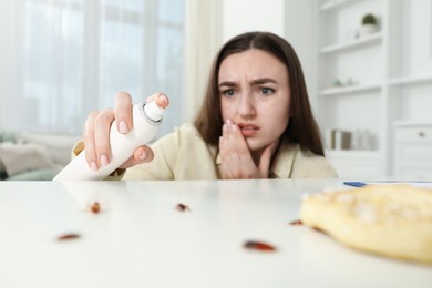 Photo of Emotional young woman spraying insecticide onto cockroaches at white table indoors, selective focus. Pest problem