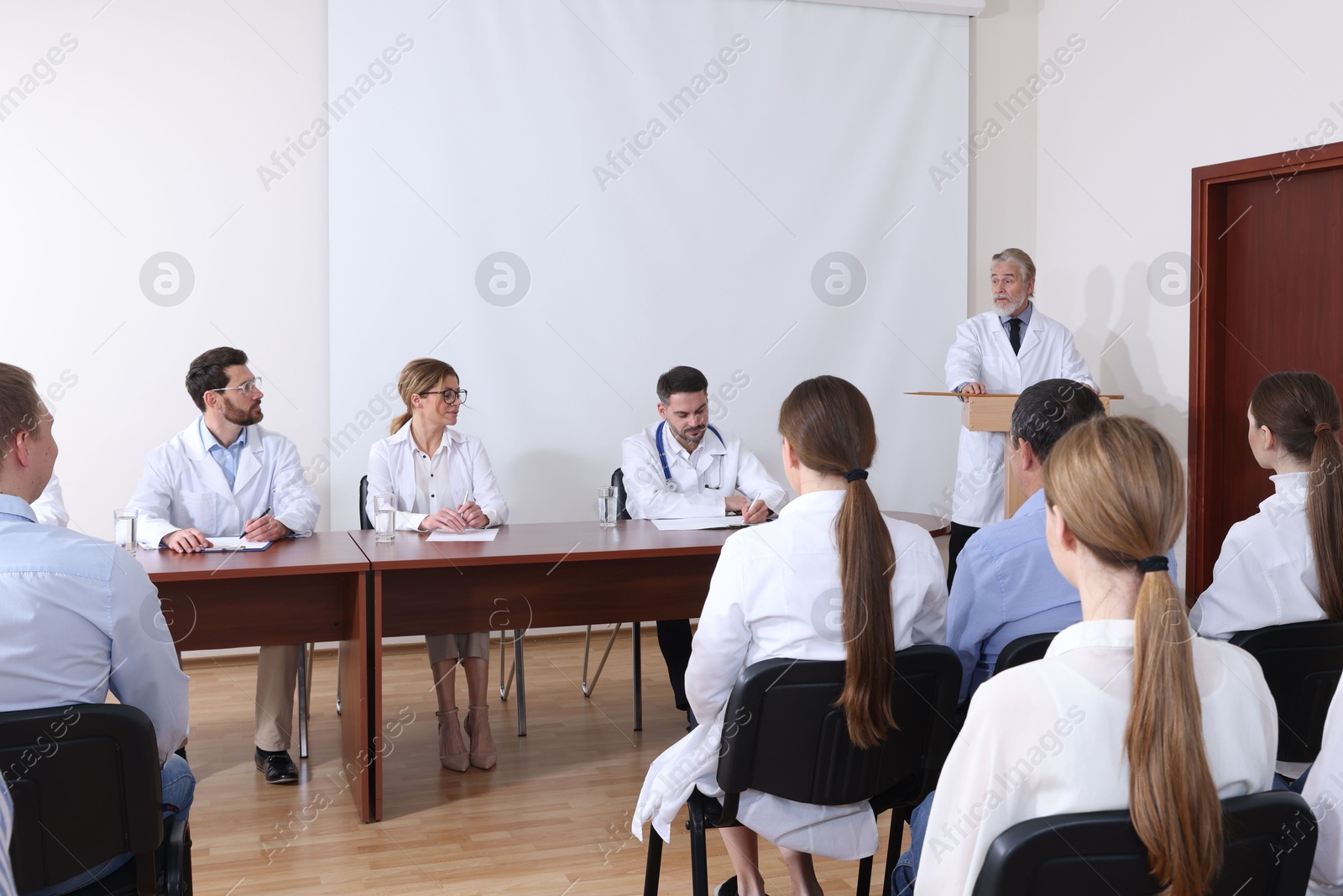 Senior doctor giving lecture in conference room with projection screen Photo of Senior doctor giving lecture in conference room with projection screen