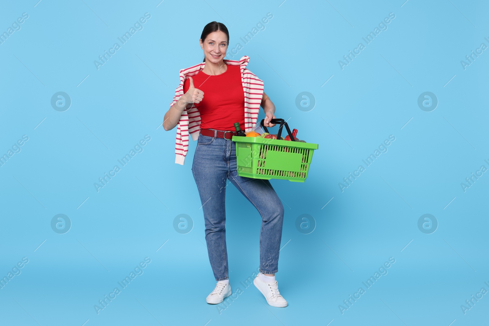 Smiling woman with shopping basket full of products showing thumbs up on light blue background Photo of Smiling woman with shopping basket full of products showing thumbs up on light blue background