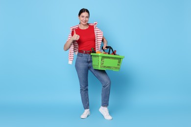 Photo of Smiling woman with shopping basket full of products showing thumbs up on light blue background