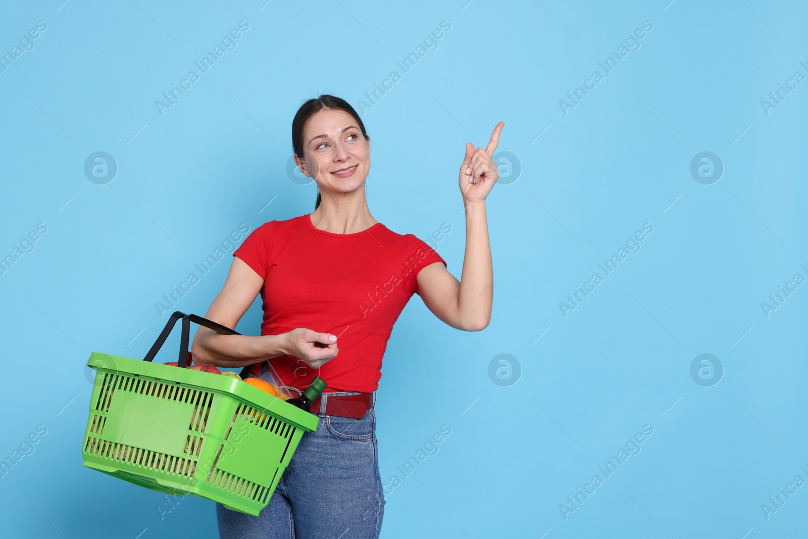 Photo of Smiling woman with shopping basket full of products pointing at something on light blue background. Space for text
