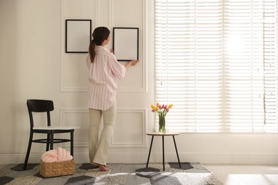 Photo of Woman hanging blank frames on white wall indoors, back view