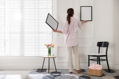 Photo of Woman hanging blank frames on white wall indoors, back view