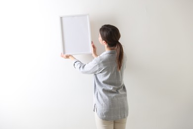 Photo of Woman hanging blank frame on white wall, back view