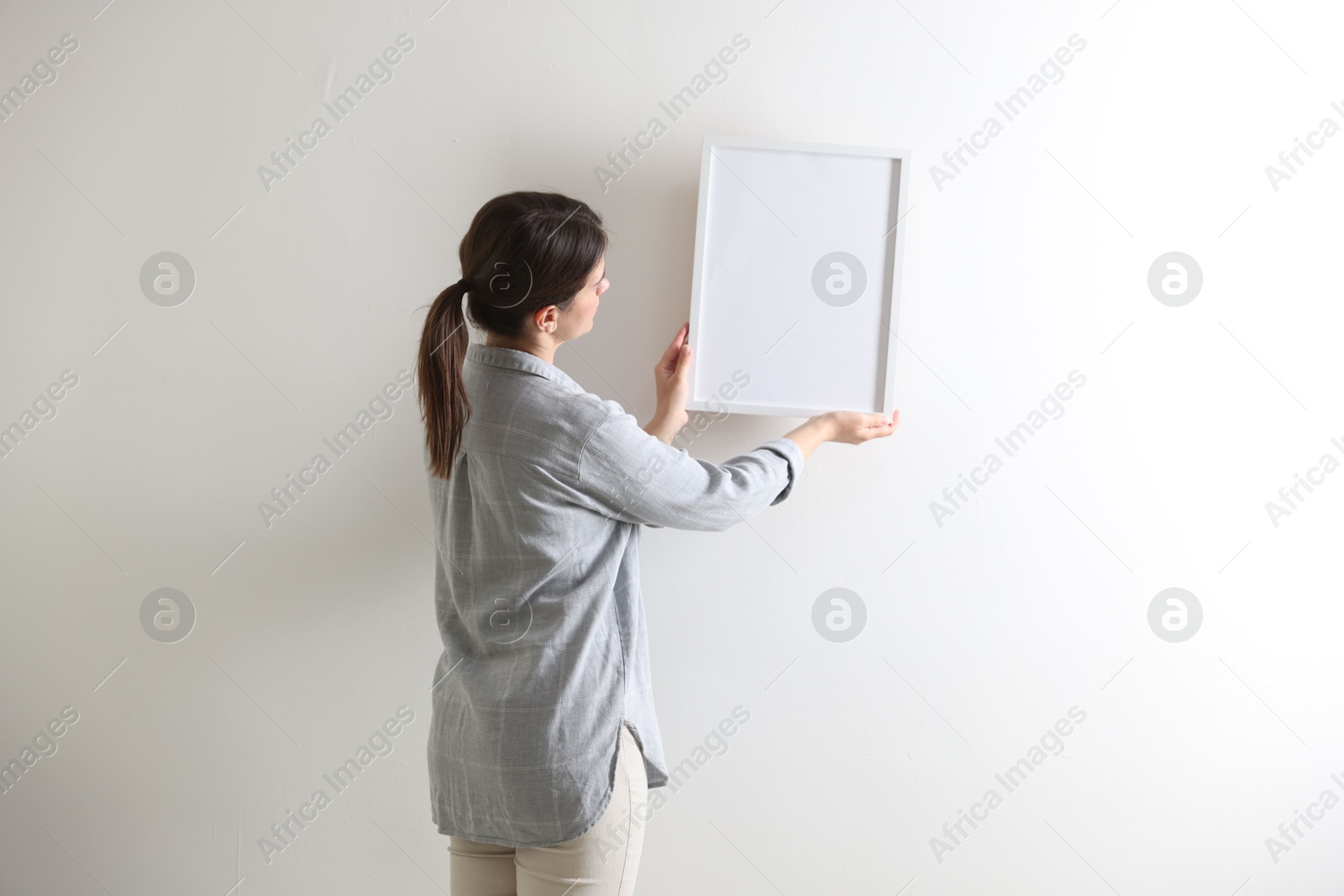 Photo of Woman hanging blank frame on white wall, back view
