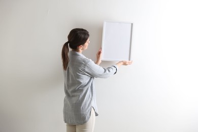 Photo of Woman hanging blank frame on white wall, back view