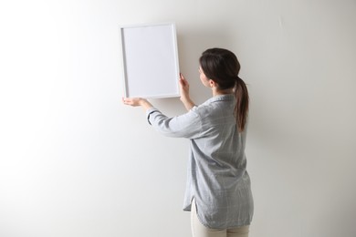 Photo of Woman hanging blank frame on white wall, back view