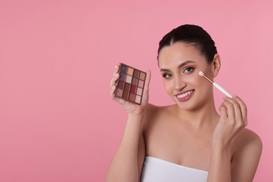 Photo of Smiling woman with eyeshadow palette and brush applying makeup on pink background. Space for text