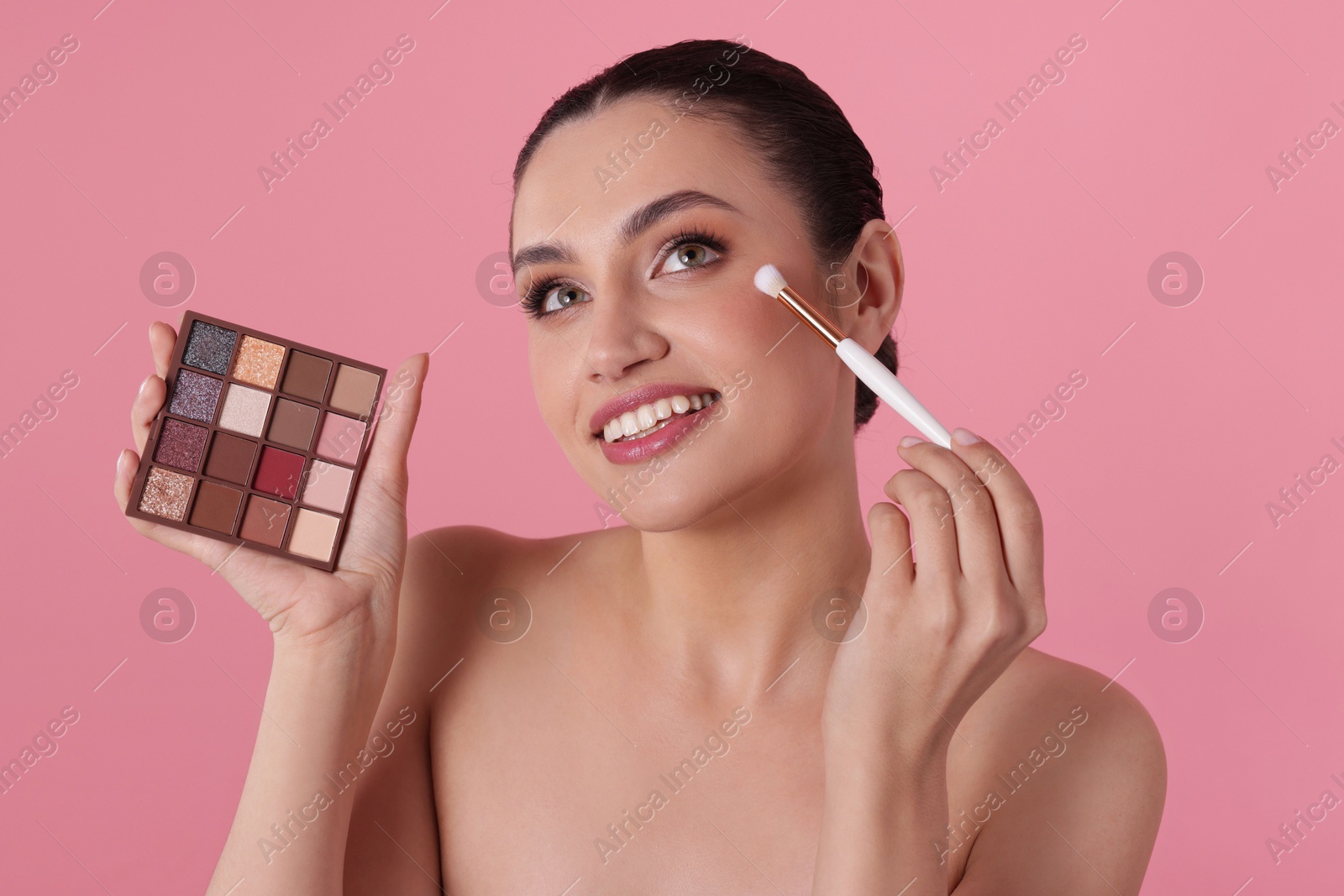Smiling woman with eyeshadow palette and brush applying makeup on pink background Photo of Smiling woman with eyeshadow palette and brush applying makeup on pink background