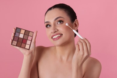 Photo of Smiling woman with eyeshadow palette and brush applying makeup on pink background