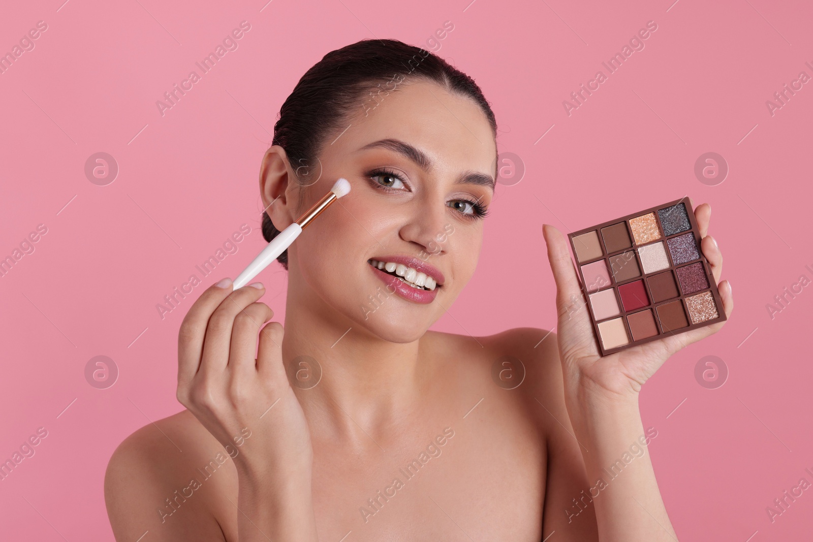 Smiling woman with eyeshadow palette and brush applying makeup on pink background Photo of Smiling woman with eyeshadow palette and brush applying makeup on pink background