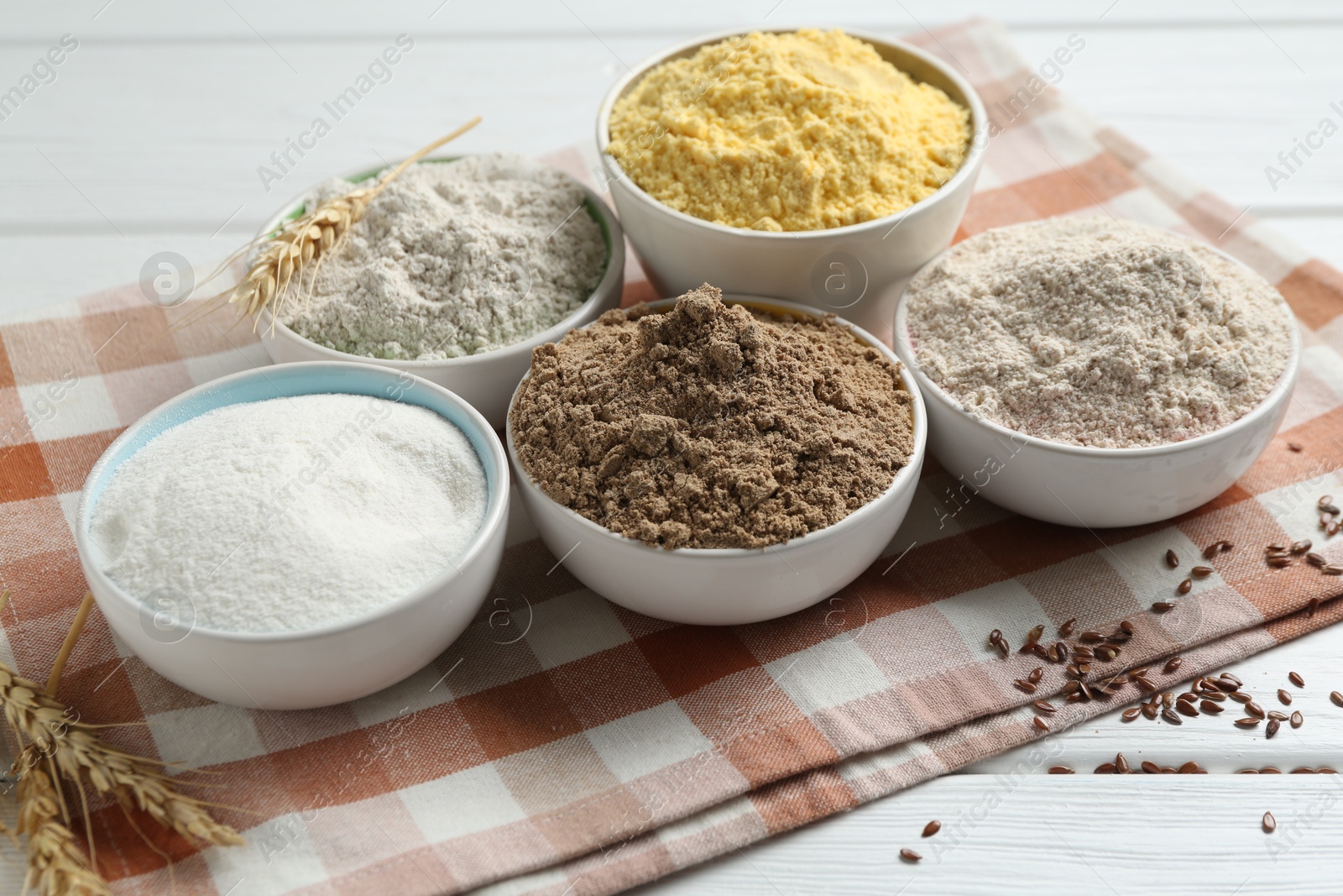 Photo of Different types of flour in bowls, spikes and flaxseeds on white wooden table, closeup