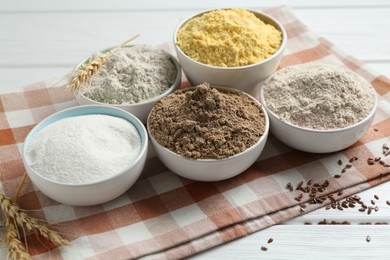 Photo of Different types of flour in bowls, spikes and flaxseeds on white wooden table, closeup