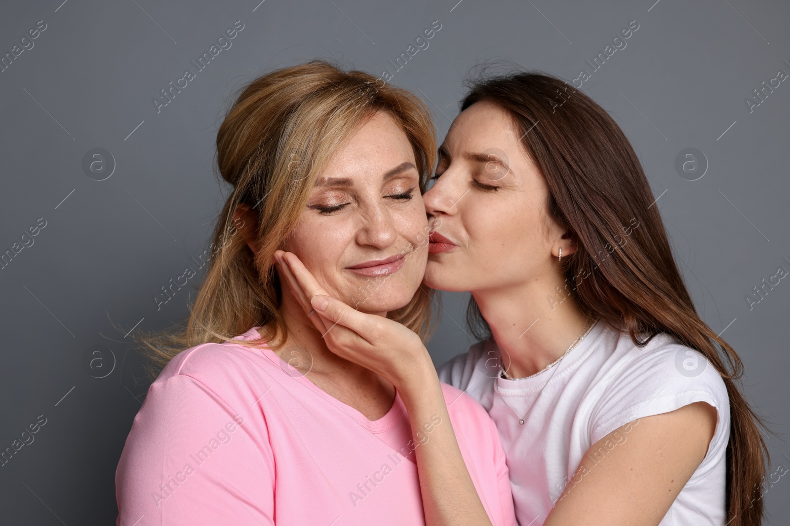 Woman kissing her mother on cheek against grey background Photo of Woman kissing her mother on cheek against grey background