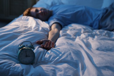 Photo of Frustrated man suffering from insomnia on bed indoors, focus on alarm clock