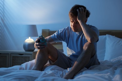 Photo of Frustrated man looking at alarm clock on bed at night. Insomnia problem