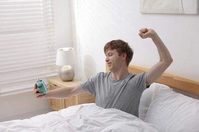 Photo of Man with alarm clock stretching in bed indoors. Good morning