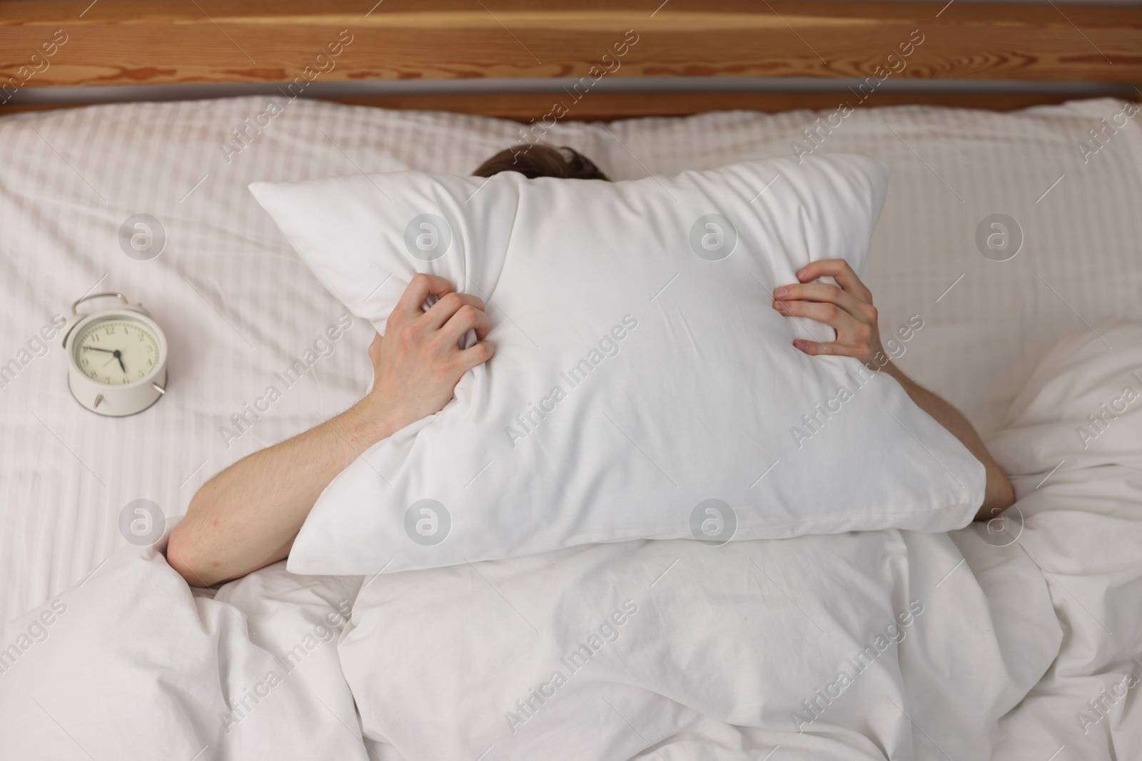 Tired man covering with pillow and alarm clock in bed indoors, above view Photo of Tired man covering with pillow and alarm clock in bed indoors, above view