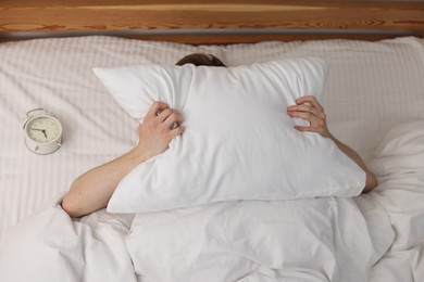 Photo of Tired man covering with pillow and alarm clock in bed indoors, above view