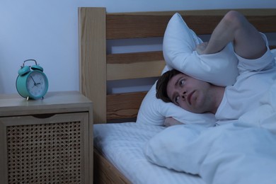 Photo of Frustrated man covering ears with pillow on bed at night. Insomnia problem