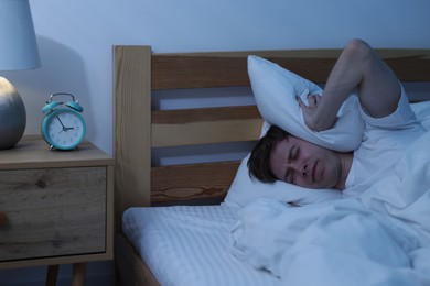 Photo of Frustrated man covering ears with pillow on bed at night. Insomnia problem