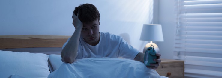 Photo of Frustrated man looking at alarm clock on bed at night. Insomnia problem