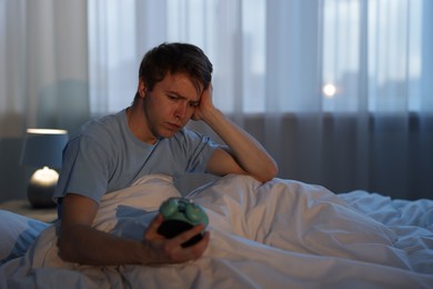 Photo of Frustrated man looking at alarm clock on bed at night. Insomnia problem