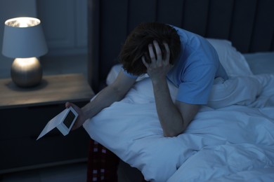 Photo of Frustrated man looking at digital alarm clock on bed at night. Insomnia problem