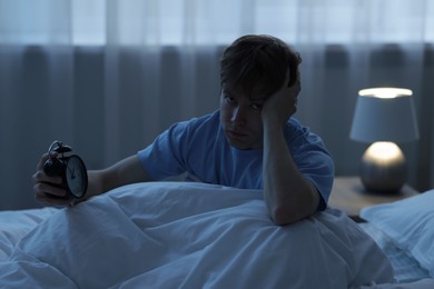 Photo of Frustrated man with alarm clock on bed at night. Insomnia problem