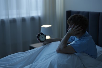 Photo of Frustrated man looking at alarm clock on bed at night. Insomnia problem