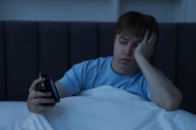 Photo of Frustrated man looking at alarm clock on bed at night. Insomnia problem