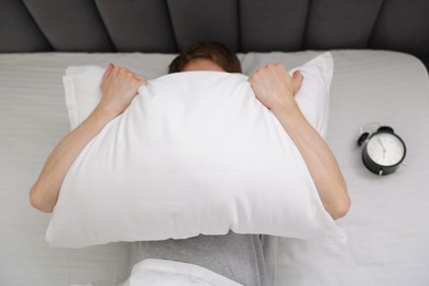 Photo of Tired man covering with pillow and alarm clock in bed indoors, above view
