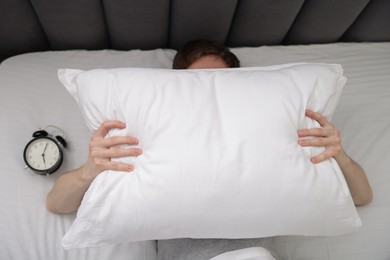 Photo of Tired man covering with pillow and alarm clock in bed indoors, top view