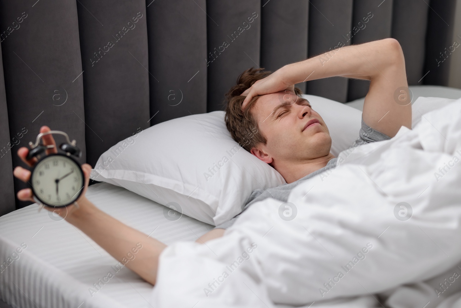 Sleepy man with alarm clock in bed indoors, selective focus Photo of Sleepy man with alarm clock in bed indoors, selective focus