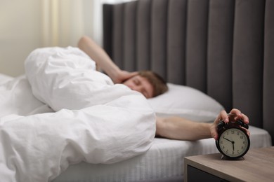Photo of Sleepy man turning off alarm clock in bed indoors, selective focus