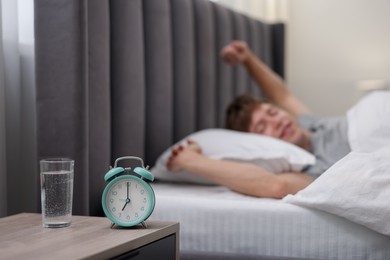Photo of Happy man awakening in bed indoors, focus on alarm clock and glass of water