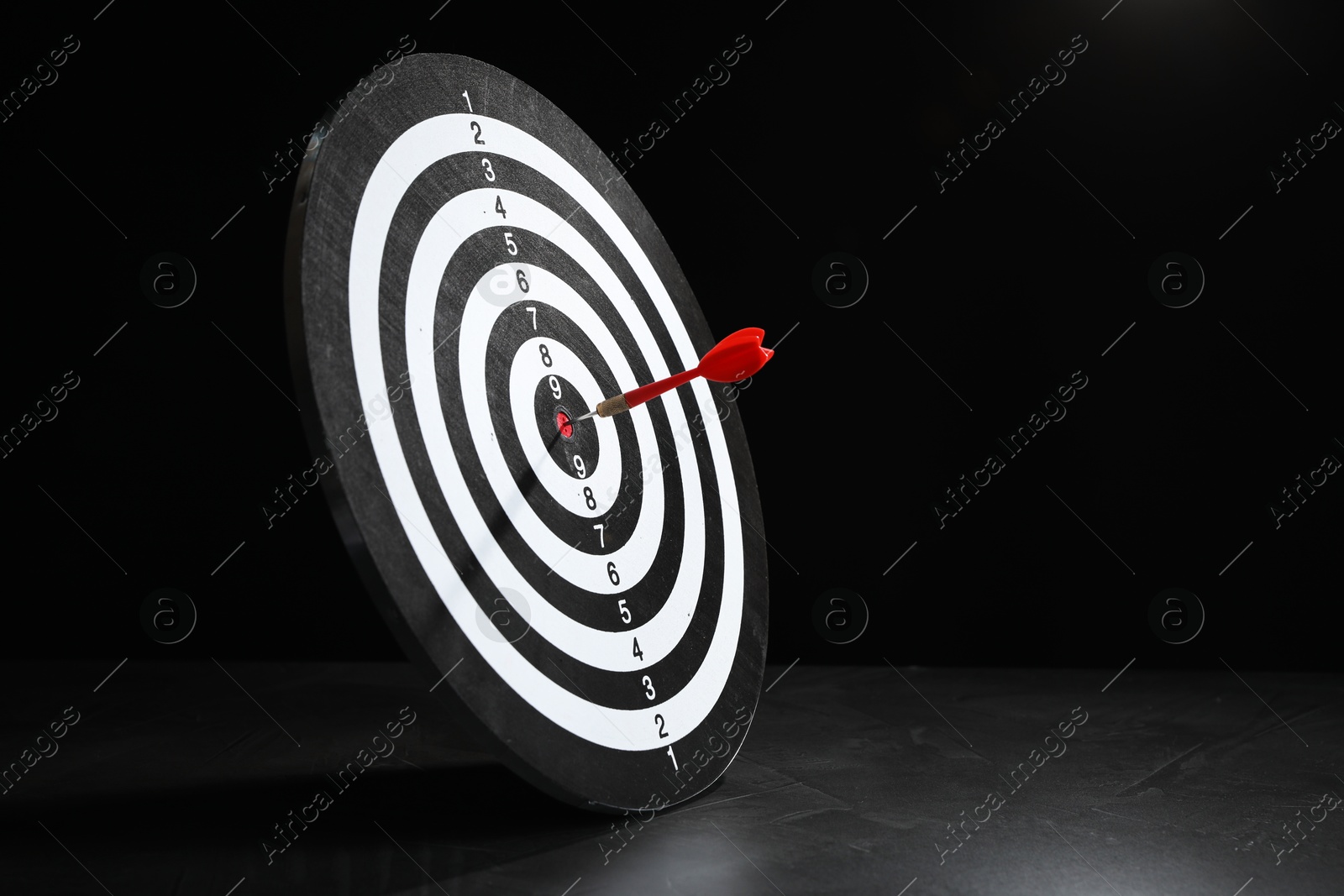 Targeting. Dart board with arrow on gray surface against black background, space for text Photo of Targeting. Dart board with arrow on gray surface against black background, space for text