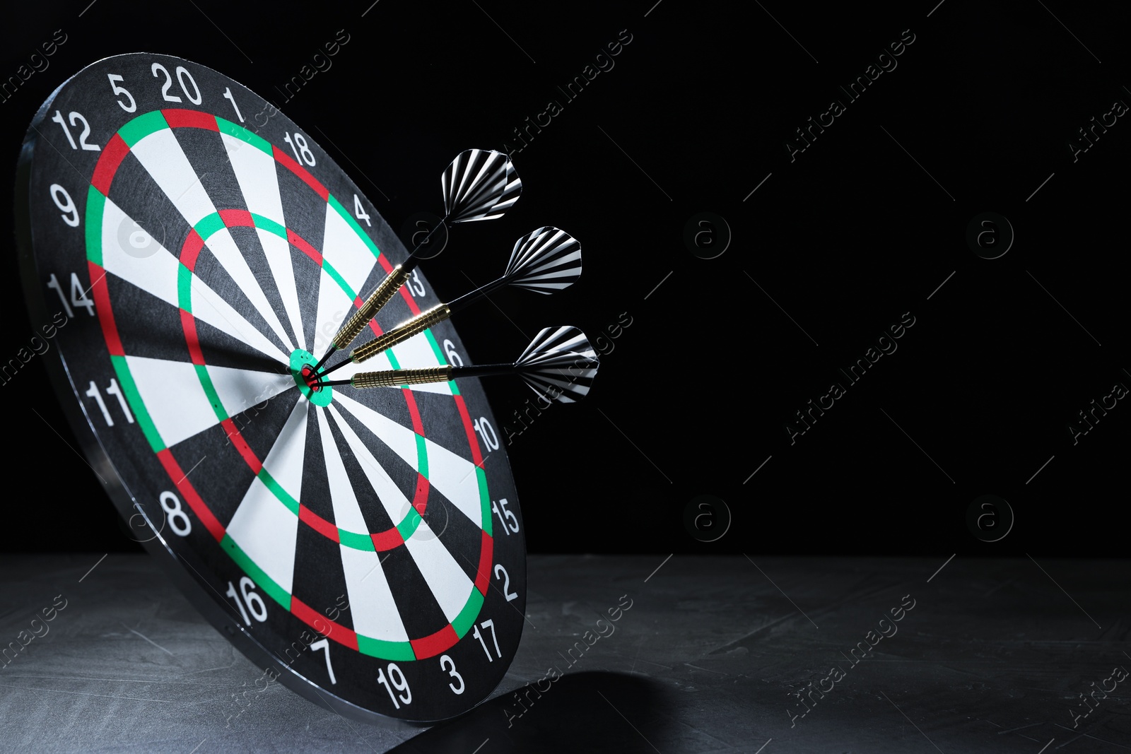 Targeting. Dart board with arrows on gray surface against black background, space for text Photo of Targeting. Dart board with arrows on gray surface against black background, space for text