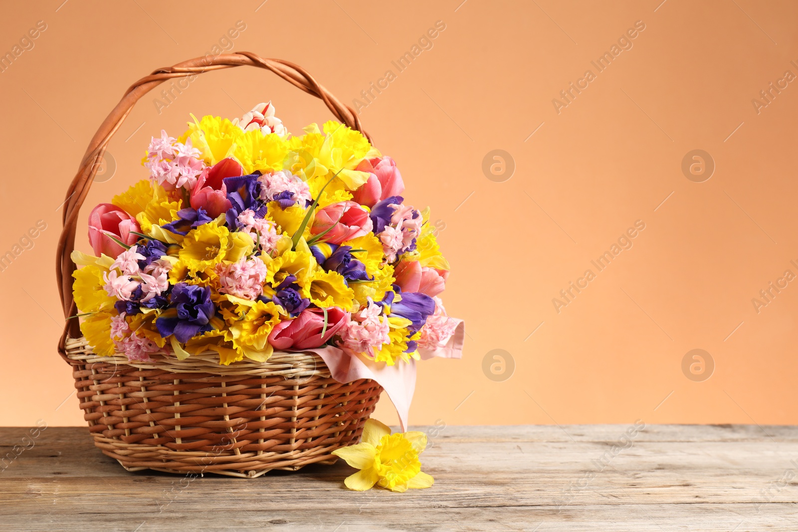 Wicker basket with beautiful flowers on wooden table against coral background. Space for text Photo of Wicker basket with beautiful flowers on wooden table against coral background. Space for text