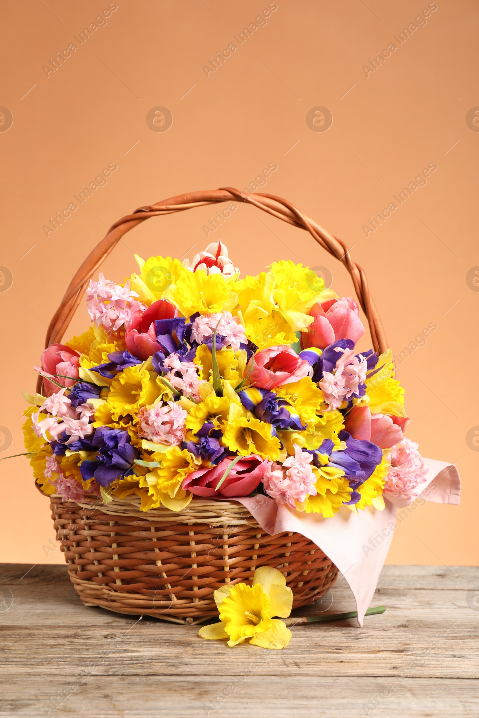 Wicker basket with beautiful flowers on wooden table against coral background Photo of Wicker basket with beautiful flowers on wooden table against coral background