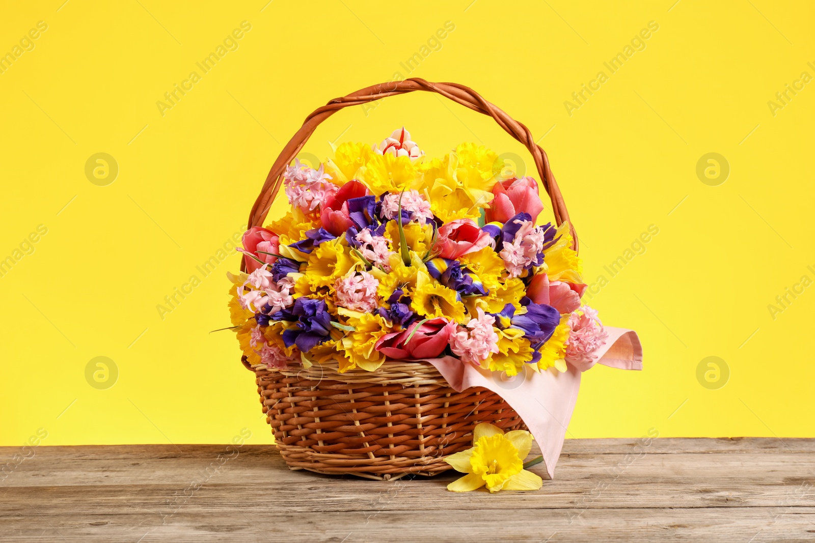 Wicker basket with beautiful flowers on wooden table against yellow background Photo of Wicker basket with beautiful flowers on wooden table against yellow background