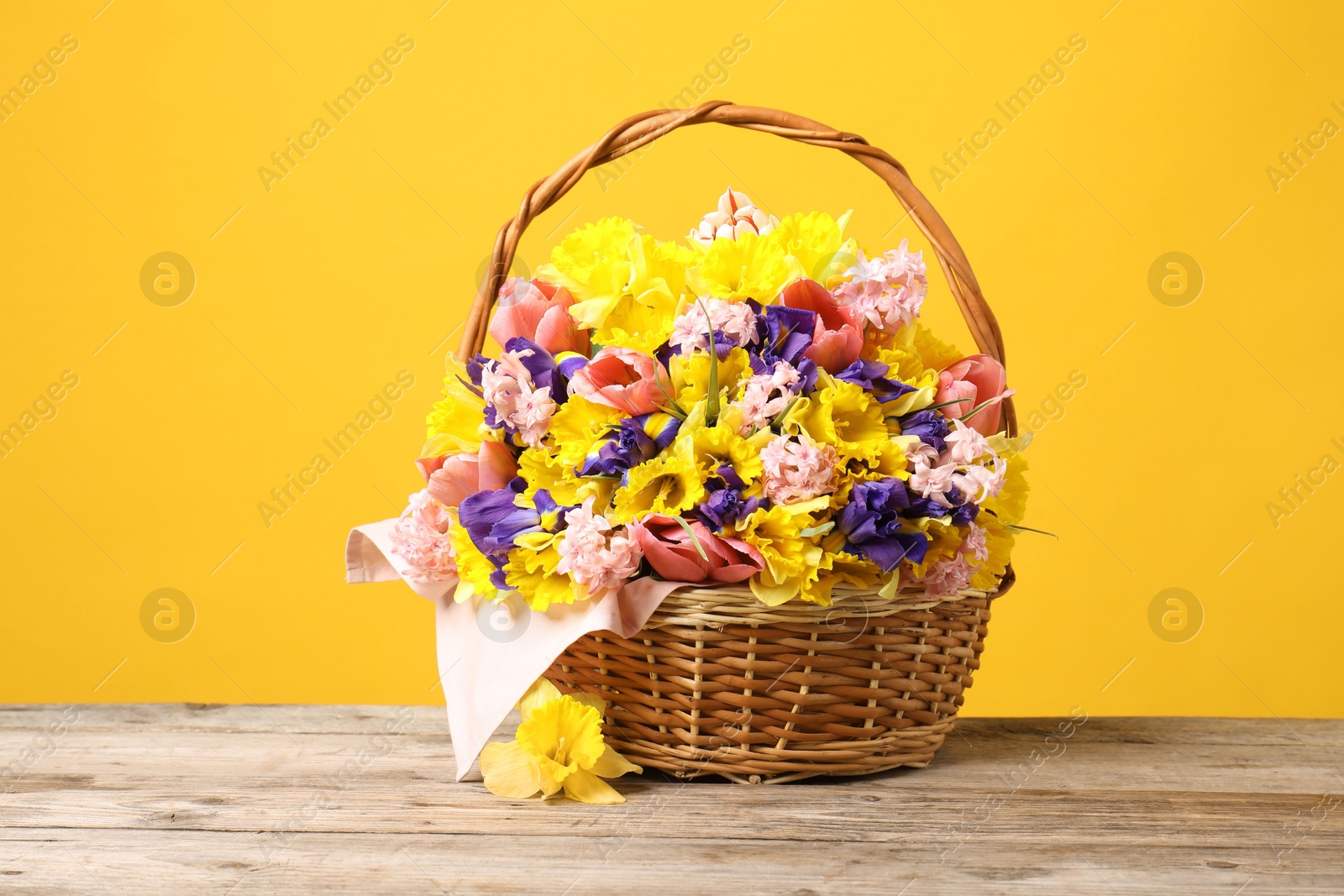 Wicker basket with beautiful flowers on wooden table against yellow background Photo of Wicker basket with beautiful flowers on wooden table against yellow background