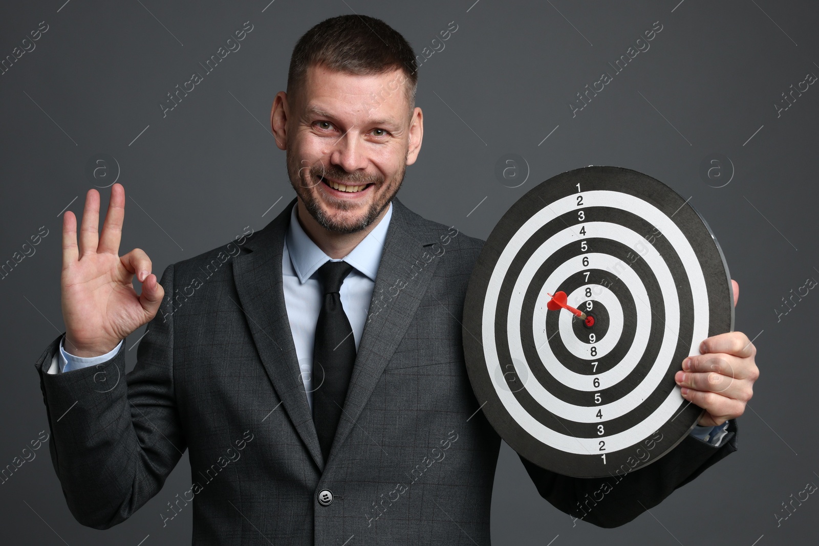 Targeting. Man with dart board and arrow showing ok gesture on grey background Photo of Targeting. Man with dart board and arrow showing ok gesture on grey background
