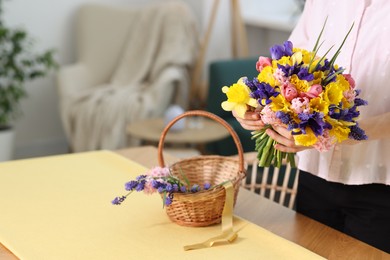 Photo of Woman with bouquet of beautiful flowers and wicker basket at table indoors, closeup