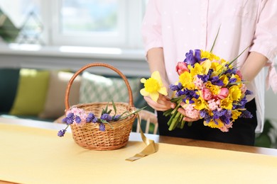 Photo of Woman with bouquet of beautiful flowers and wicker basket at table indoors, closeup