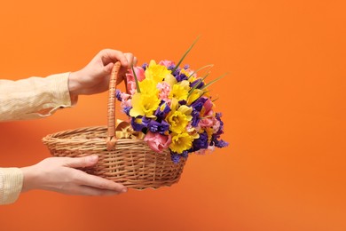 Photo of Woman holding wicker basket with beautiful flowers on orange background, closeup. Space for text