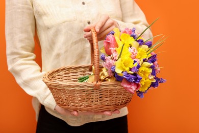 Photo of Woman holding wicker basket with beautiful flowers on orange background, closeup