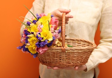 Photo of Woman holding wicker basket with beautiful flowers on orange background, closeup